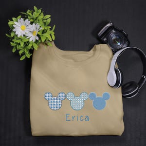 May include: A tan sweatshirt with three blue and white Mickey Mouse heads embroidered on the front. The name "Erica" is embroidered below the Mickey Mouse heads.