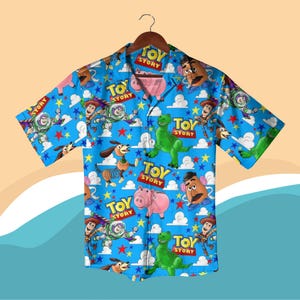 K&ouml;nnte beinhalten: Ein kurz&auml;rmeliges Button-Down-Hemd mit einem leuchtend blauen Hintergrund, der mit Figuren und den Worten "Toy Story" gemustert ist. Das Design umfasst Woody, Buzz Lightyear, Rex, Hamm und Mr. Potato Head.
