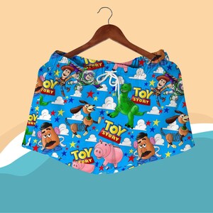 K&ouml;nnte beinhalten: Blaue Shorts mit einem sich wiederholenden Muster von Toy Story-Charakteren, darunter Woody, Buzz Lightyear und Rex. Die Shorts haben einen wei&szlig;en Kordelzug und h&auml;ngen an einem Holzb&uuml;gel. Die Worte "Toy Story" sind auf die Shorts gedruckt.