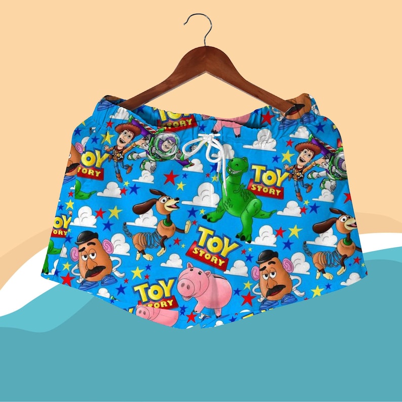 K&ouml;nnte beinhalten: Blaue Shorts mit einem sich wiederholenden Muster von Toy Story-Charakteren, darunter Woody, Buzz Lightyear und Rex. Die Shorts haben einen wei&szlig;en Kordelzug und h&auml;ngen an einem Holzb&uuml;gel. Die Worte "Toy Story" sind auf die Shorts gedruckt.