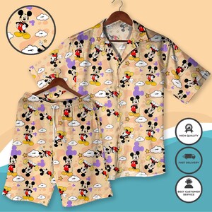 May include: A beige button-down shirt and matching shorts set. The set features a repeating pattern of Mickey Mouse, clouds, and stars in red, black, yellow, and purple. The shirt has short sleeves and a collar. The shorts have a drawstring waist.