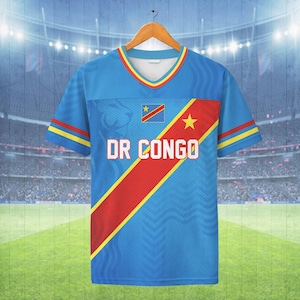 Op de afbeelding: Een blauw T-shirt met V-hals, een diagonale rode streep met een gele ster, de vlag van de Democratische Republiek Congo en de tekst DR CONGO in witte letters. De mouwen hebben rode en gele strepen en een leeuwengrafiek is zichtbaar.