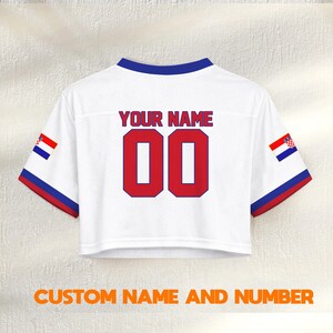 K&ouml;nnte beinhalten: Wei&szlig;es Crop-Top-Trikot mit blauen und roten Akzenten. Der Text "YOUR NAME" steht &uuml;ber der roten Nummer "00". Enth&auml;lt kroatische Flaggen-Details auf den &Auml;rmeln. Der Text "CUSTOM NAME AND NUMBER" steht unten.