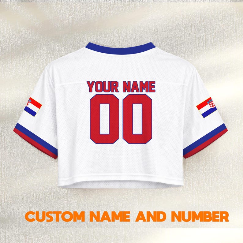 K&ouml;nnte beinhalten: Wei&szlig;es Crop-Top-Trikot mit blauen und roten Akzenten. Der Text "YOUR NAME" steht &uuml;ber der roten Nummer "00". Enth&auml;lt kroatische Flaggen-Details auf den &Auml;rmeln. Der Text "CUSTOM NAME AND NUMBER" steht unten.