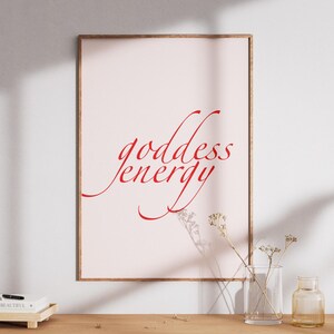 May include: A framed print with the words "goddess energy" in red script. The print is displayed on a light pink background. The frame is a light brown color. The print is displayed on a wooden cabinet with a small tray, a vase, and a small bottle.