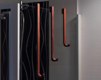 May include: Three wooden hooks hang from a glass shower door. The hooks are a reddish-brown color and have a simple, curved design.