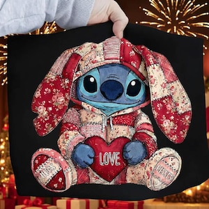 Puede incluir: Un bolso tote negro con un personaje de dibujos animados azul que lleva una sudadera con capucha de retazos y sostiene un corazón rojo con la palabra "LOVE". El personaje tiene orejas largas estampadas con tela floral roja. El bolso tiene "HUGS" y "KISSES" en los lados.
