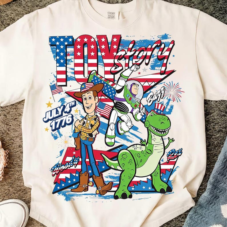 May include: White t-shirt featuring a graphic of Woody, Buzz Lightyear, and Rex from Toy Story. The design incorporates American flag elements and the text "Toy Story" and "July 4th 1776".