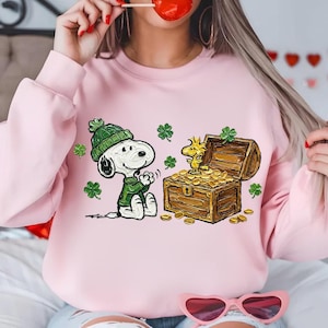 Puede incluir: Sudadera rosa con un diseño del Día de San Patricio. Presenta a Snoopy con gorro y suéter verdes, un cofre del tesoro lleno de monedas de oro y Woodstock. Tréboles verdes están dispersos.
