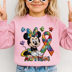 Puede incluir: Sudadera rosa claro con Minnie Mouse, un lazo de rompecabezas y una cinta de piezas de rompecabezas. La palabra "AUTISMO" está escrita con piezas de rompecabezas de colores. El diseño está centrado.