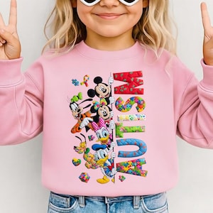 Puede incluir: Sudadera rosa con los personajes de Disney Mickey Mouse, Minnie Mouse, Goofy, Daisy Duck y Donald Duck. La palabra "AUTISMO" está escrita con letras de piezas de rompecabezas de colores. La sudadera tiene mangas largas.