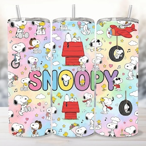 Puede incluir: Tres vasos de acero inoxidable con un diseño colorido de Snoopy. El diseño presenta a Snoopy en varias poses, junto con Woodstock, sobre un fondo de arcoíris pastel. La palabra "Snoopy" se muestra en letras grandes y coloridas.