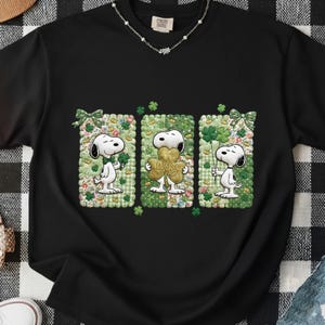 Puede incluir: Camiseta negra con personajes de Snoopy sosteniendo tréboles y un trébol dorado "Lucky". El diseño incluye patrones florales y de cuadros verdes y blancos, con lazos verdes. Un diseño gráfico temático del Día de San Patricio.