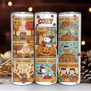 Puede incluir: Un vaso blanco con paneles de cómic que ilustran escenas otoñales. Los paneles muestran personajes con calabazas y hojas, con texto como "APPLE PICKKIN!", "COZY", "LEAF PILE FUN!", "POP CORNS!", "AUTUMN TALE!" y "PUMPKIN-TASTIC!".