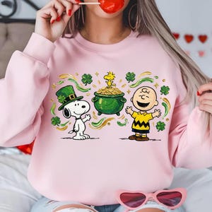 Puede incluir: Sudadera rosa claro con un diseño de dibujos animados de Snoopy con un sombrero de duende verde y Charlie Brown con una olla de oro y tréboles. El diseño es de estilo caricatura.