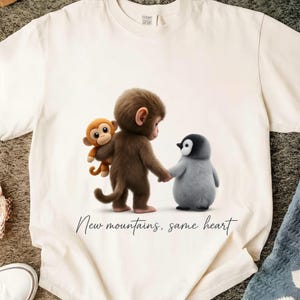 May include: Cream-colored t-shirt featuring a whimsical illustration of a brown monkey holding hands with a gray penguin, with a smaller orange monkey on the monkey's back. The text "New mountains, same heart" is printed below the image.