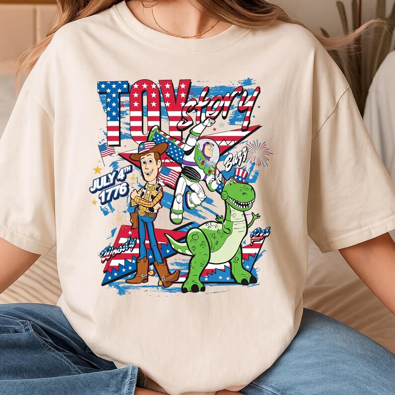 May include: Cream-colored t-shirt featuring a graphic of Toy Story characters Woody, Buzz Lightyear, and Rex, with patriotic red, white, and blue accents. The text "Toy Story" and "July 4th 1776" are also displayed.