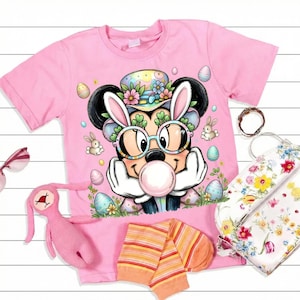 Puede incluir: Camiseta rosa con un personaje de dibujos animados de Mickey Mouse con orejas de conejo, gafas y una burbuja de chicle. El diseño incluye huevos de Pascua y conejitos. También se muestra un conejito de peluche rosa, calcetines y un bolso floral.