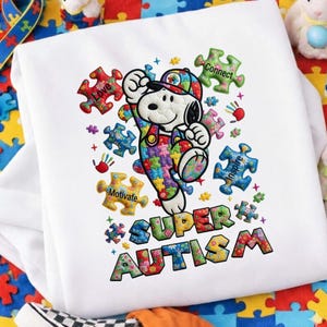 Puede incluir: Sudadera blanca con Snoopy vestido con un atuendo de piezas de rompecabezas de colores, con las palabras "Super Autism" en un diseño floral. Piezas de rompecabezas con palabras como "Love", "Connect" e "Inspire" rodean a Snoopy.