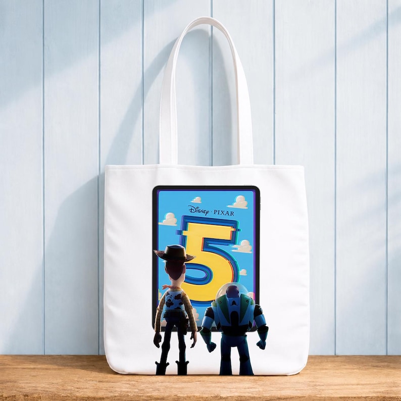 Pu&ograve; includere: Una borsa tote bianca con una stampa grafica del logo Disney Pixar Toy Story 5, con i personaggi Woody e Buzz Lightyear in piedi davanti ad esso. Il design mostra il grande numero cinque giallo su uno sfondo di cielo azzurro.
