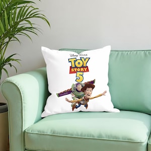Pu&ograve; includere: Un cuscino quadrato bianco con il logo Disney Pixar Toy Story 5 in testo blu, giallo e rosso. Sotto il logo, i personaggi Woody e Buzz Lightyear sono raffigurati in una posa dinamica, con Buzz che vola e Woody che tende la mano.