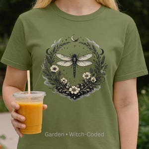 Model wearing Comfort Colors 1717 t-shirt in Washed Denim, standing in a lush garden. Dragonfly Sigil visible on chest, surrounded by botanical elements and crescent moon. Relaxed fit, vintage washed cotton. Witchy, soft dark aesthetic for intuitive, nature-loving women.