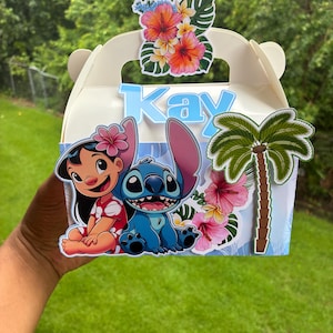 May include: A white cardboard gable box featuring a cutout of Lilo and Stitch, a palm tree, and tropical hibiscus flowers. The name Kay is displayed in light blue 3D lettering on the front. The box has a handle and a decorative floral topper.