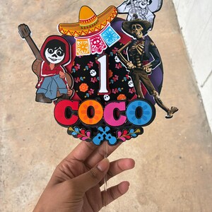 May include: A colorful Coco-themed cake topper featuring characters and elements from the movie. The topper includes the word "COCO" in vibrant letters, the number "1", and images of a sombrero, guitar, and skeleton figures. The design is held by a clear stick.