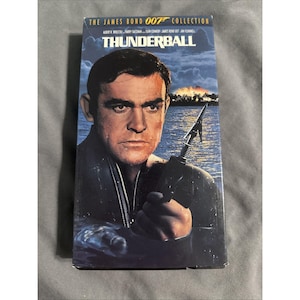 May include: A VHS tape of the James Bond 007 Collection film Thunderball. The cover features a portrait of Sean Connery holding a speargun, with a dark blue water and sky background, and the title Thunderball printed in bold white letters.