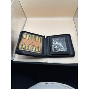May include: A black zippered travel case containing a portable backgammon board with green and yellow triangular points on a tan surface. The right side features a clear plastic pocket holding a collection of metal game pieces and two dice.