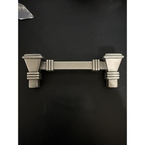 May include: A silver-toned cabinet pull with a rectangular center and decorative end pieces. The ends have a stepped design, and the center has vertical lines. This hardware is suitable for drawers or cabinets.