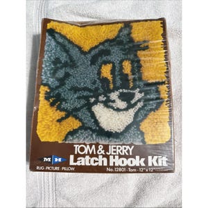 May include: A Tom & Jerry latch hook kit featuring a cartoon image of Tom the cat. The kit includes materials for a rug, picture, or pillow, measuring 12 inches by 12 inches. The design showcases Tom's face with yellow, gray, and white colors.