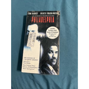 May include: Sealed VHS tape of the movie "Philadelphia" starring Tom Hanks and Denzel Washington. The cover features images of the actors and the film's title in red. Includes the music video "Streets of Philadelphia" and details of the 1993 Academy Awards.