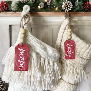 May include: Two white knitted Christmas stockings with wooden bead garlands and red wooden tags. The tags are personalized with the names "Mom" and "Hailey".