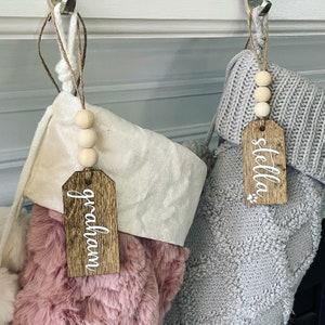 May include: Two Christmas stockings with personalized wooden tags. The stocking on the left is pink and fluffy, with a tag that says "graham". The stocking on the right is gray and knitted, with a tag that says "stella".