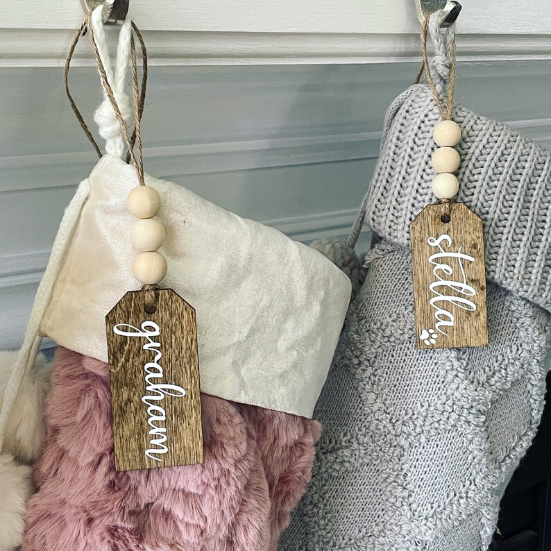 May include: Two Christmas stockings with personalized wooden tags. The stocking on the left is pink and fluffy, with a tag that says "graham". The stocking on the right is gray and knitted, with a tag that says "stella".