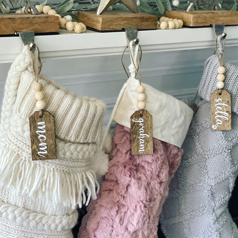 May include: Three Christmas stockings with wooden name tags. The stockings are white, pink, and gray. The name tags read "Mom", "Graham", and "Stella".