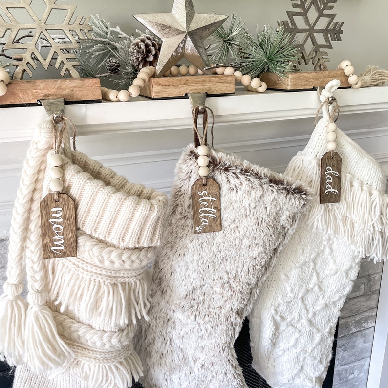 May include: Three white Christmas stockings with wooden tags that say "mom", "stella", and "dad" hanging on a white fireplace mantel.