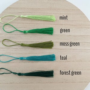 May include: Five different colored tassels, from top to bottom: mint, green, moss green, teal, and forest green. The tassels are arranged in a row on a wooden surface.