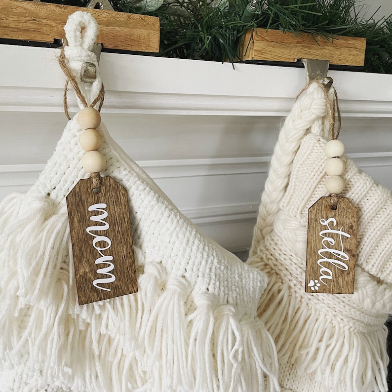 May include: Two white crocheted stockings with wooden tags that say "Mom" and "Stella". The stockings are hung on a white fireplace mantel with wooden hooks.