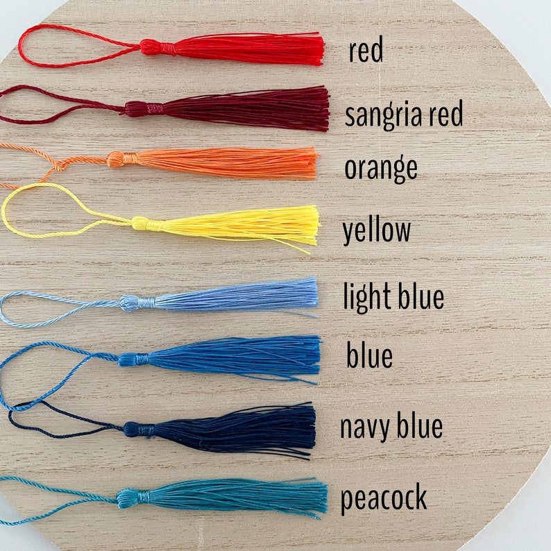 May include: A set of 9 colorful tassels in various shades of red, orange, yellow, blue, and green. The tassels are arranged in a row on a wooden surface. The colors are labeled with their names: red, sangria red, orange, yellow, light blue, blue, navy blue, and peacock.