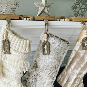 May include: Three Christmas stockings with personalized wooden tags hanging on a white fireplace mantel. The stockings are white with a fringe, brown faux fur, and gold velvet. The tags read "Brady", "Mom", and "Opie".