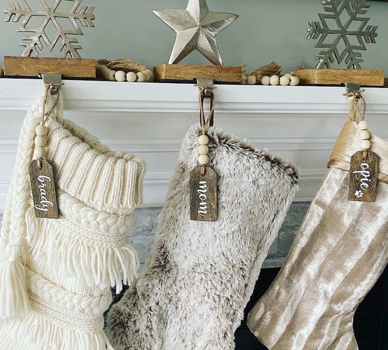 May include: Three Christmas stockings with personalized wooden tags hanging on a white fireplace mantel. The stockings are white with a fringe, brown faux fur, and gold velvet. The tags read "Brady", "Mom", and "Opie".