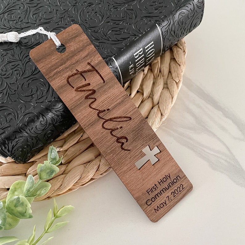 May include: A wooden bookmark with the name "Emilia" engraved on it. It also features a silver cross and the text "First Holy Communion May 7, 2022".