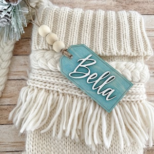 May include: A white knitted stocking with a blue wooden tag that says "Bella". The tag is attached to the stocking with twine and wooden beads.
