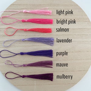 May include: Seven different colored tassels, from light pink to mulberry, arranged in a row on a wooden surface. The tassels are all the same size and shape, and they are made of a soft, silky material.