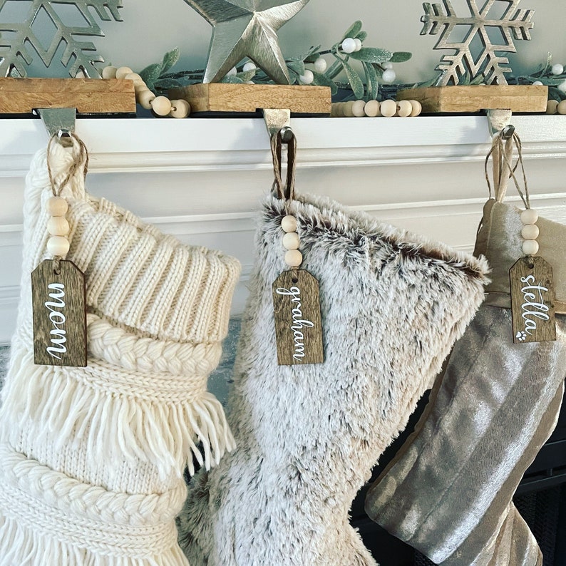 May include: Three white, brown, and beige Christmas stockings with wooden tags that say "mom", "graham", and "stella" hanging on a white mantel with a silver star and snowflake ornament.
