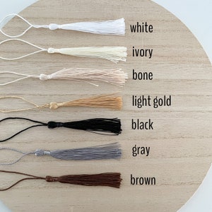 May include: Seven different colored tassels with cords, arranged in a row on a wooden surface. The tassels are white, ivory, bone, light gold, black, gray, and brown. The colors are labeled below each tassel.