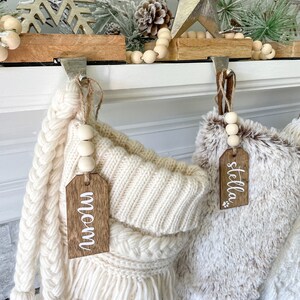 May include: Two white knitted stockings with wooden tags that say "mom" and "stella". The stockings are hung on a white mantel with wooden hooks and a garland of wooden beads.