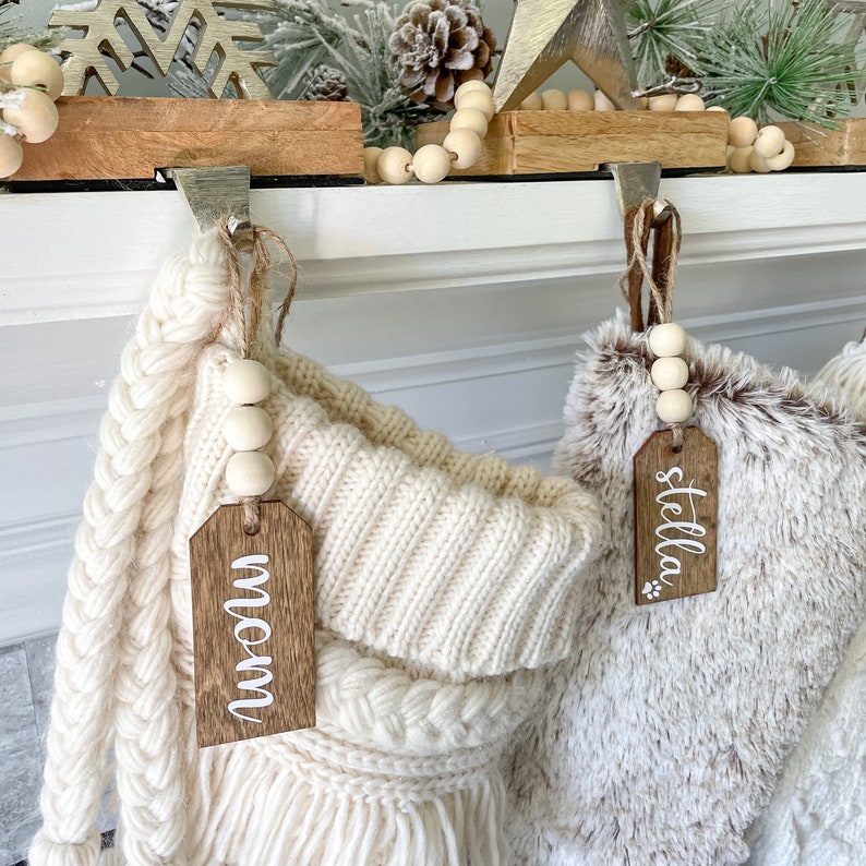 May include: Two white knitted stockings with wooden tags that say "mom" and "stella". The stockings are hung on a white mantel with wooden hooks and a garland of wooden beads.